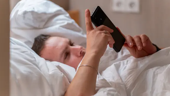 Ein junger Mann liegt im Bett und schaut auf sein Smartphone, während er nicht einschlafen kann und durch Nachrichten oder Unterhaltung vom Schlaf abgehalten wird