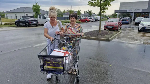 Eine blonde Frau schiebt mit einer älteren Frau einen Einkaufswagen. 