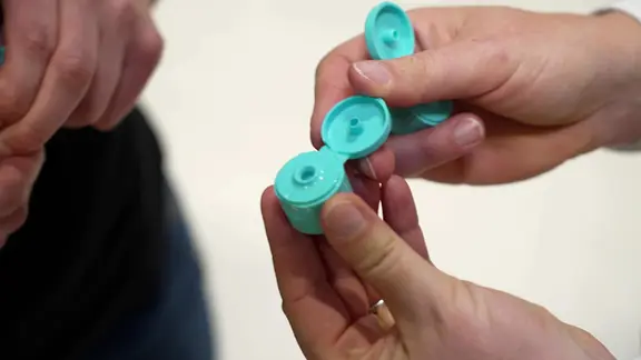 Zwei Hände halten zwei türkisfarbigen Plastikklappverschlüsse für Kosmetiktuben oder -flaschen.