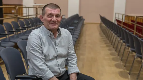Steffen Gerisch: Mann im Hemd und dunkler Hose sitzt auf einem Stuhl einer leeren Sitzreihe, er hat kurzrasierte dunkle Haare und lächelt 