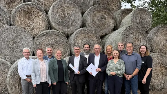 Eine Gruppe von Menschen, die einen Förderbescheid erhält, steht vor riesigen, aufeinandergeschichteten Ballen aus getrocknetem Hanf.