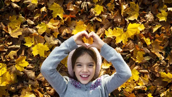 Ein Kind zeigt mit den Händen ein Herz vor Herbstblättern.