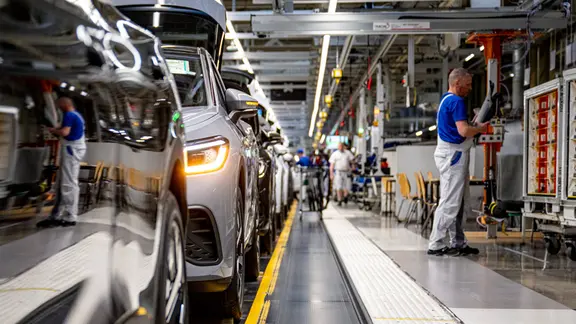 Mitarbeiter in blauem T-Shirt und weißer Latzhose neben mehreren neuen Volkswagen hintereinander bei der Endkontrolle