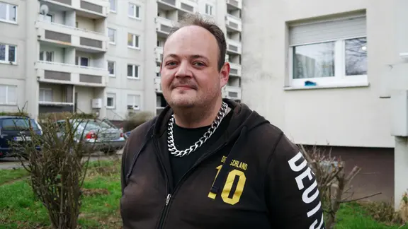 Ein Mann mit Trainingsjacke und silberner Kette steht vor einigen Wohnhäusern.
