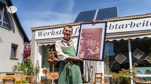 Ein Mann in grüner Schürze steht in einem Korb vor einer Werkstatt und schaut freundlich in dei Kamera.