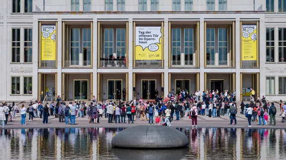 Eine Menge Menschen steht vor dem Opernhaus, ein Gebäude mit Terasse und zahlreichen Fenstern.