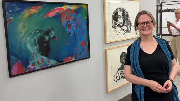 April Eisman vor Arbeiten von Angela Hampel: Eine lächelnde Frau mit blondem Zopf und Brille in schwarzer Kleidung und langem blauen Schal vor einer Wand mit gerahmter Kunst
