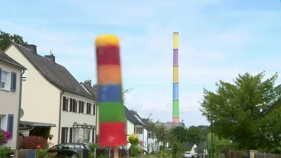 Ein buntes Stieleis im Vordergrund hat dieselben Farben wie die bunte Esse von Chemnitz, die im Hintergrund zu sehen ist. 