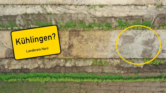 Luftaufnahme einer archäologischen Ausgrabungsfläche mit parallelen, unterschiedlich gefärbten Erdstreifen und Grabungsschnitten. Ein gelb umkreister Bereich rechts markiert rechteckige Bodenverfärbungen, vermutlich Siedlungsspuren. Eingeblendet ist ein stilisiertes gelbes Ortsschild mit der Aufschrift „Kühlingen" mit Fragezeigchen und dem Zusatz „Landkreis Harz".