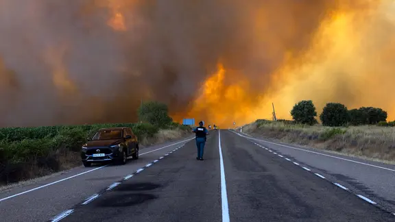 Ein massiver Waldbrand mit orangefarbenen Flammen und dichtem, gelb-braunem Rauch, der eine Landstraße überzieht. Ein dunkles Auto steht am Straßenrand, eine Person in dunkler Uniform mit reflektierendem Streifen geht auf der Mittellinie der Straße in Richtung der Rauchwolken. Rechts und links der Straße stehen Bäume und Sträucher vor landwirtschaftlichen Feldern.
