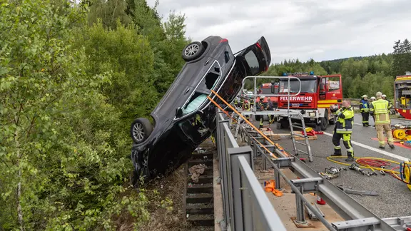 Beschädigtes schwarzes Auto hängt nach Unfall an Autobahnbrücke und wird von Feuerwehrleuten geborgen