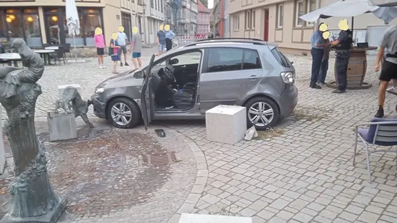 Ein Auto mit offener Fahrertür steht auf einem gepflasterten Platz vor Häusern und Stehtischen, neben einer kaputten Statue