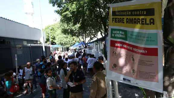 Menschen stehen in Schlange für Impfungen. Im Vordergrund ein Schild, auf dem "Impfung gegen Gelbfieber" auf portugiesisch steht.