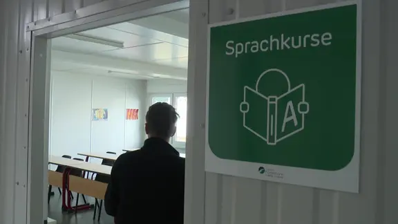 Eine Person steht in einem Eingang zum Klassenraum. An der Tür hängt ein Schild mit der Aufschrift: Sprachkurse. 