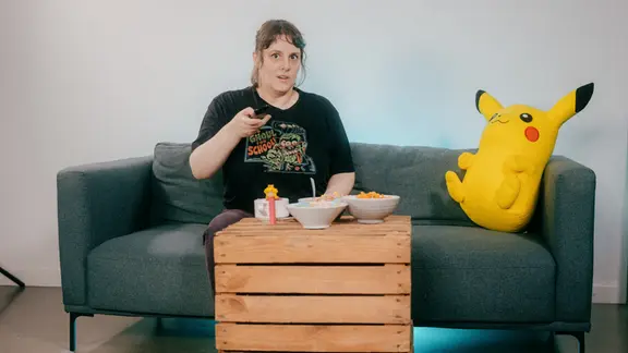 Eine Frau sitzt auf einem Sofa mit einer großen Pickachu-Figur und zappt mit der Fernbedienung. Vor ihr ein Couchtisch mit diversen Snacks.