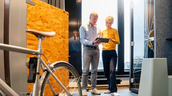 Mann und Frau stehen in einem hellen Büro mit Holztrennwand und betrachten gemeinsam ein Tablet; ein Fahrrad ist im Vordergrund unscharf zu sehen.
