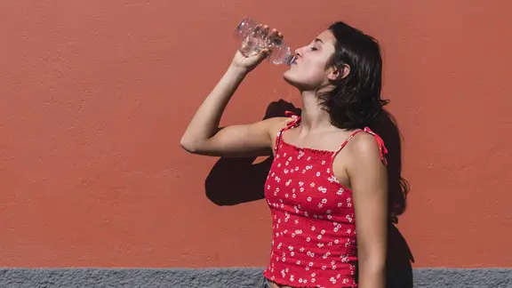 Eine junge Frau mit langen, dunklen Haaren trinkt aus einer transparenten Plastikwasserflasche.