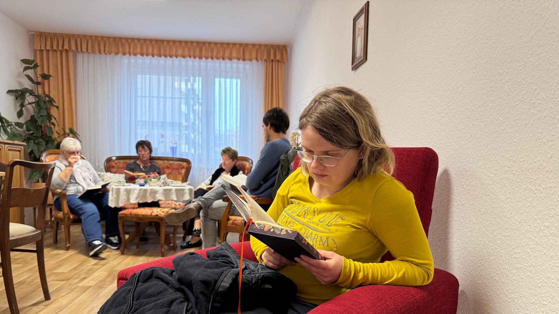 Eine junge Frau mit einem gelben Pullover sitzt in einem roten Sessel und liest leise ein Buch. | Vanessa Clobes