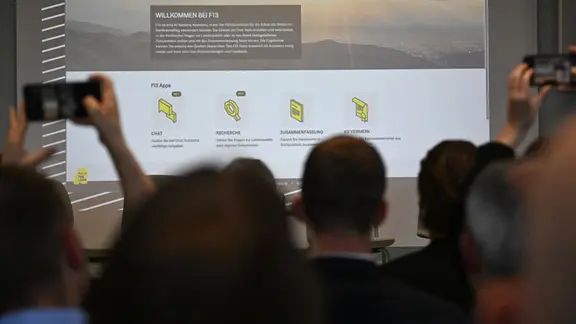 Bei einer Präsentation des neuen KI-Assistenzsystems F13 ist auf eine Leinwand die Benutzoberfläche projiziert und Zuschauer im Vordergrund machen mit Smartphones Fotos davon