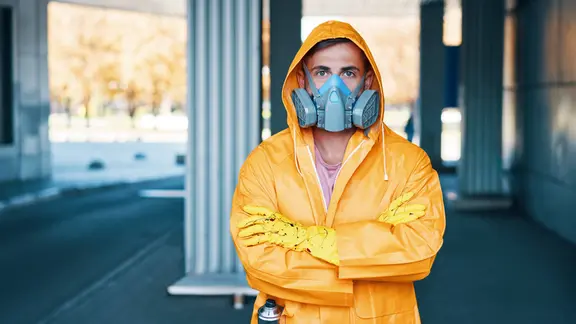 Nahaufnahme eines jungen Mannes mit Atemschutzmaske und gelbem Regenmantel, der auf der Straße posiert und in die Kamera schaut.