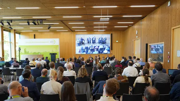 In einem mit Holz verkleidetem Saal sitzen zahlreiche Männer und Frauen und schauen auf ein Podium.