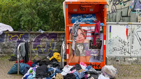 Aufgerissene Müllsäcke und wild umhergeworfene Kleidung liegen vor einem Altkleidercontainer. 
