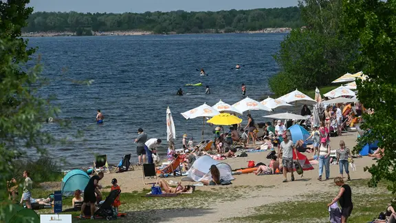 Ausflugsgäste am Badestrand schauen auf die Segelboote auf dem Cospudener See. Bei warmen Temperaturen genießen zahlreiche Besucher die entspannte Atmosphäre und nutzen das sonnige Wetter für Erholung, Aktivitäten im Freien und gemeinsame Zeit an diesem beliebten Ausflugsziel.. (zu dpa: «Wetterbilanz: Frühling hat Sachsen mit viel Sonne verwöhnt»)