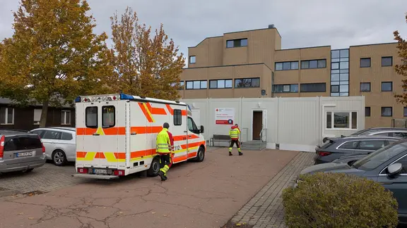 Ein Krankenwagen und zwei Sanitäter vor einem Containergebäude.