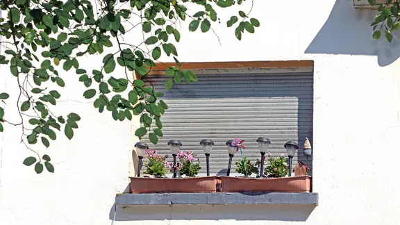 Gleißende Sonne, die während einer Hitzewelle mit voller Wucht auf eine Gebäudefassade trifft und die Fenster durch Aufheizen durch Rollladen geschützt werden. Die Blumenkästen aus Kunststoff auf dem Fensterbrett haben sich durch die Hitze verformt.