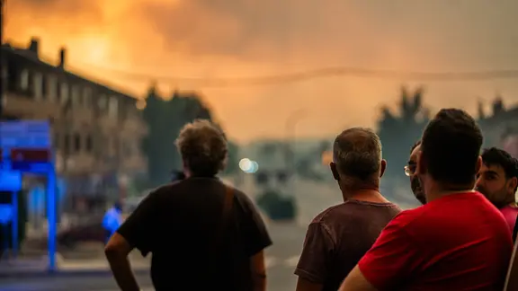 Mehrere Menschen stehen auf einer Straße mit dem Rücken zur Kamera und beobachten eine große Rauch- und Feuerwolke am Abendhimmel über einer Häuserzeile.”
