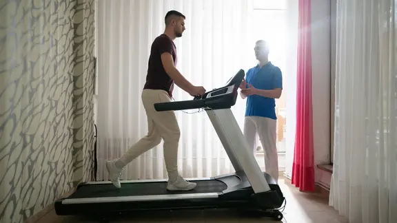 Ein Mann unterzieht sich einer physikalischen Therapie mit Hilfe eines Trainers auf einem Laufband in einem Haus.