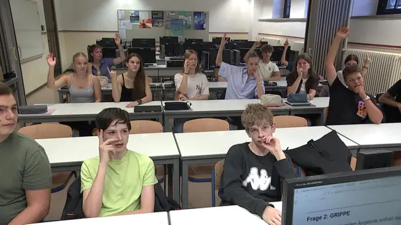 Schülerinnen und Schüler sitzen im Unterricht an Tischen und melden sich. 