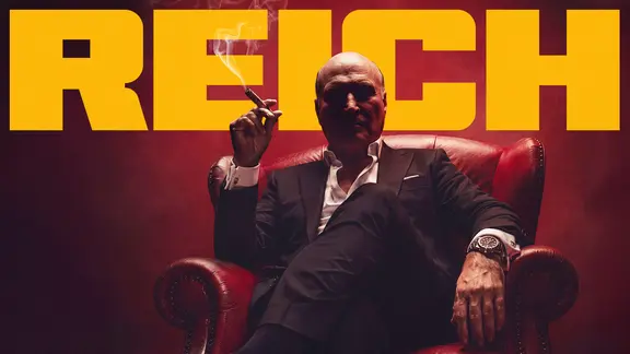 Mann mit Zigarre sitzt auf einem Sessel und blickt auf uns herab. Im Hintergrund der Titel "Reich".