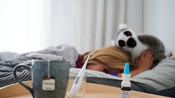 Fieberthermometer, Nasenspray und eine Tasse Tee stehen vor einem Bett, in dem ein Kind mit Kuscheltier liegt