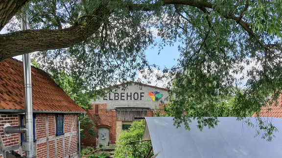 Häuser im Grünen hinter einer Wiese und einem Baum. Auf einem Haus steht "Elbehof" mit buntem Symbol dahinter