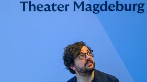 Junger Mann mit rundlichem Gesicht, Vollbart und dunklen Haaren und Brille mit dunklem Rand sitzt im Anzug vor einer blauen Wand, er blickt hoch zum Schriftzug Theater Magdeburg
