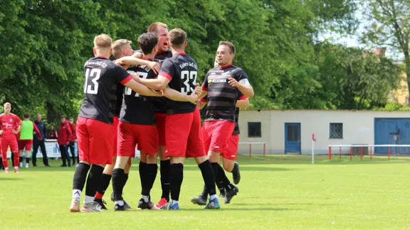 Fußballspieler der Kickers JuS 03 in schwarz-roten Trikots jubeln in einer engen Gruppenumarmung auf dem Rasenplatz, im Hintergrund Bäume und ein Vereinsgebäude mit blauen Türen.