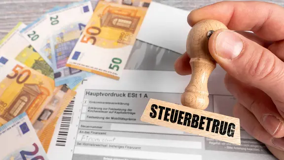 FOTOMONTAGE, Ein Stempel mit der Aufschrift Steuerbetrug wird über einer Einkommensteuererklärung gehalten. Euro-Banknoten liegen daneben und symbolisieren illegale Steuervergehen, Steuerhinterziehung und wirtschaftskriminelle Handlungen im Zusammenhang mit Finanzen.