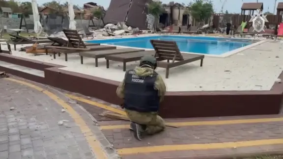 Ein Soldat hockt, vor ihm ein Poolbecken und leere Pool-Liegen. Im Hintergrund sind zerstörte Gebäude zu sehen.