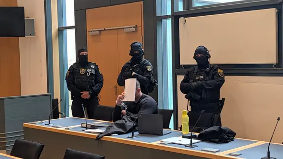 Ein Mann sitzt an einem Tisch und verdeckt sein Gesicht. Hinter ihm stehen drei vermummte Polizisten.