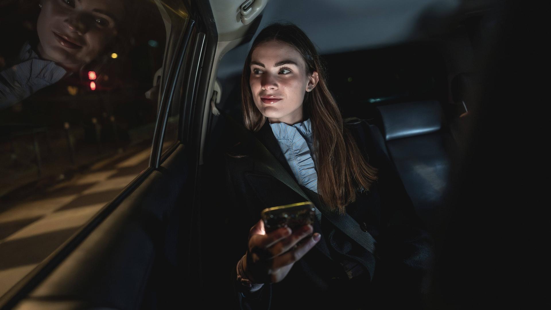 Junge Geschäftsfrau pendelt nachts mit dem Auto, sitzt auf dem Rücksitz und schaut aus dem Fenster, während sie ein Smartphone in der Hand hält. |  IMAGO / imagebroker