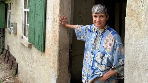 Artist in Residence in Kriebitzsch: Autorin Jennifer de Negri, Frau mit kurzen grauen Haaren und blauem Retro-Männerhemd lehnt in Tür eines alten Hauses mit grünen Fensterläden