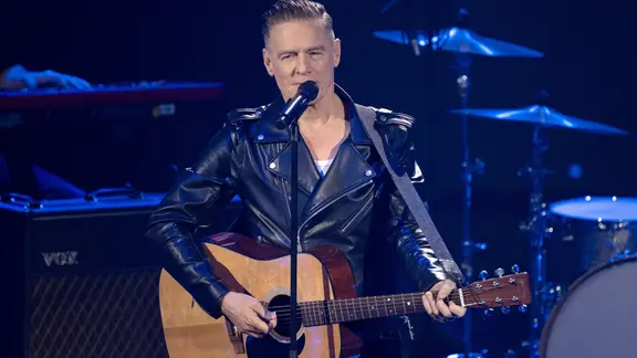 Musiker Byran Adams in schwarzer Lederjacke mit Gitarre auf einer Bühne. 
