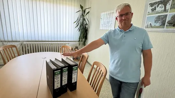 Ein Mann in einem hellblauen Polo steht neben einem Tisch auf dem Aktenordner stehen. 