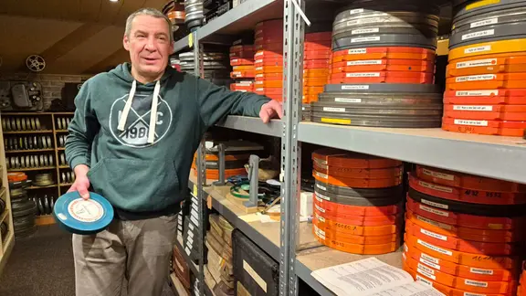 Ein Mann häleine Filmrolle in der Hand. um in herum befinden sich noch sehr viele weitere Filmrollen in Regalen gestapelt. 