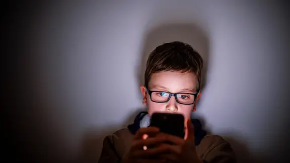 Ein Junge sitzt in einem dunklen Raum und wird von einem Smartphone beleuchtet.