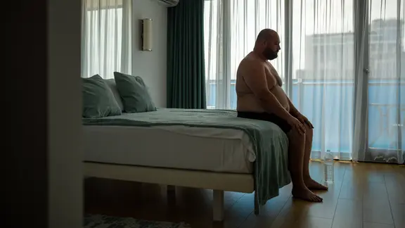 Mann sitzt oberkörperfrei in einer abgedunkelten Wohnung