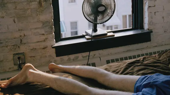 Künstlerischer Ausschnitt: Person in Boxershorts liegt auf einem Bett vor einem geöffneten Altbau-Fenster mit Backsteinwänden, davor läuft ein schwarzer Standventilator auf der Fensterbank, im Hintergrund Blick auf gegenüberliegende Stadthäuser.