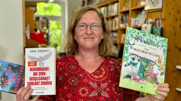 Eine Frau zeigt zwei Bücher in die Kamera und lächelt, im Hintergrund Bücherregale.