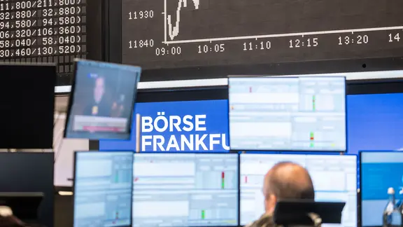 Ein Mann sitzt vor vielen Monitoren, im Hintergrund der Verlauf des DAX.